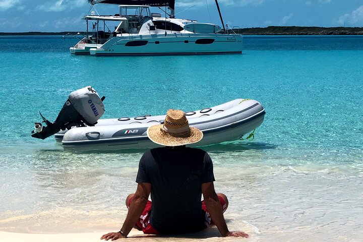 a man wearing a bucket hat while seating in the white sands in the beach in Bahamas with a small and big boat at the water
