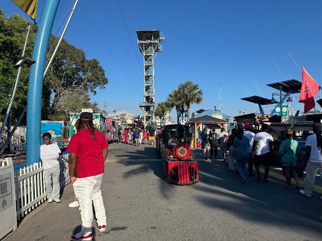A vibrant boardwalk scene in Destin, Florida, featuring a small red tourist train moving down a street lined with visitors. People stroll past food trucks and waterfront shops under a clear blue sky, with a zipline tower and palm trees in the background adding to the festive, beachside atmosphere.