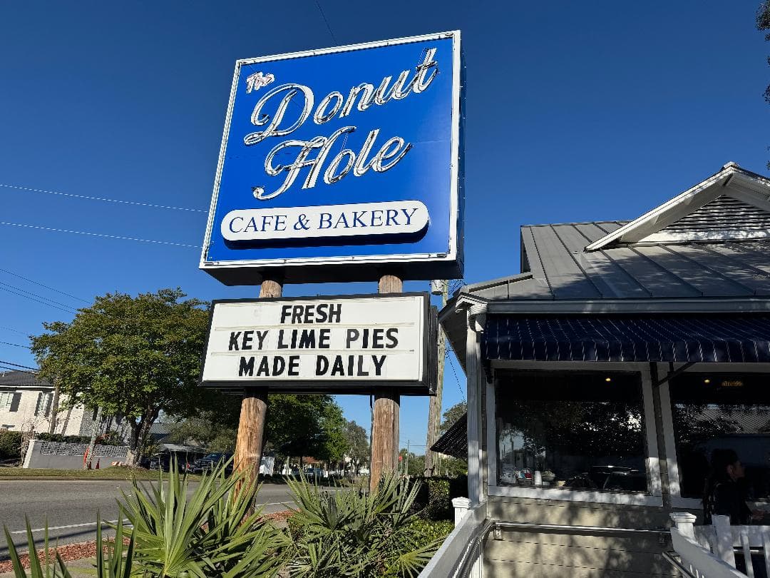 A bright blue sign for The Donut Hole Café & Bakery stands above a white marquee advertising “Fresh Key Lime Pies Made Daily.” The building has a metal roof and blue-striped awning, with a small patio area where patrons can be seen through the windows.