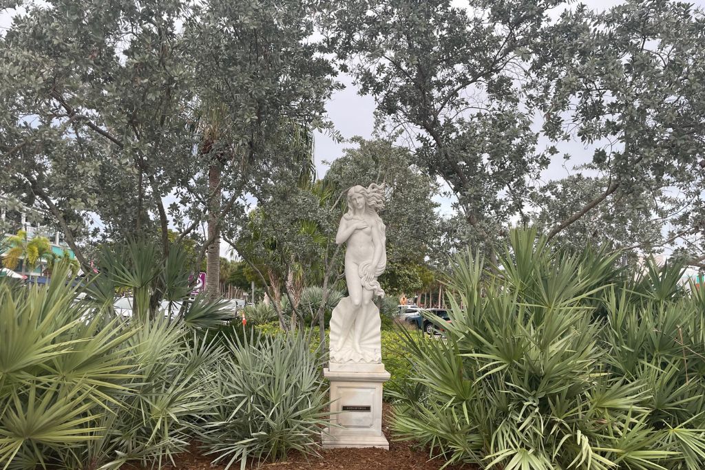 White marble statue of a nude woman with long flowing hair standing on a pedestal, surrounded by dense green foliage and silver-leafed trees. The statue appears to be inspired by classical Greco-Roman art and is set outdoors near a street with cars partially visible in the background.