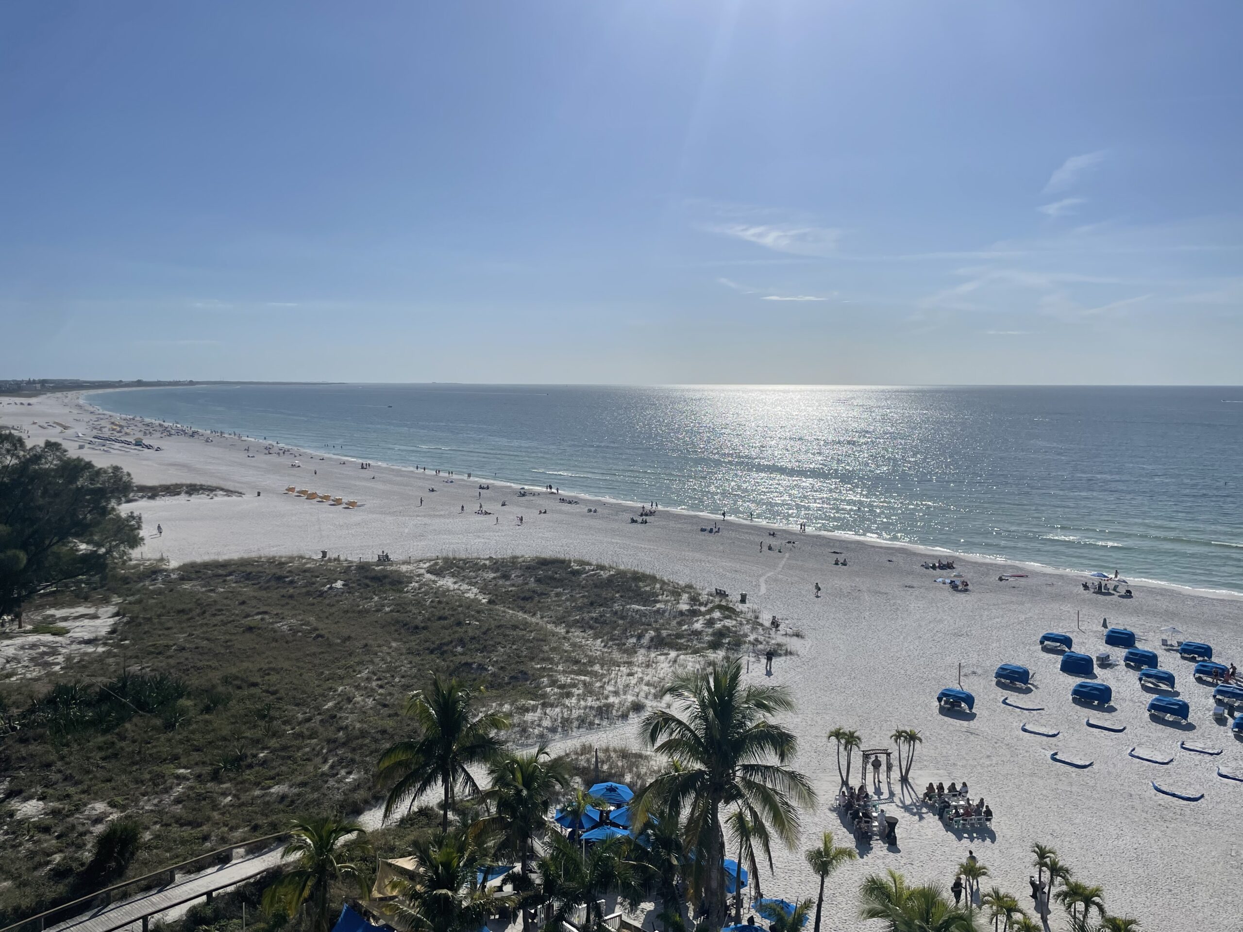 Aerial view of a sunny beach in St. Petersburg, Florida, with white sand stretching along the coastline and the Gulf of Mexico shimmering under a clear blue sky. Scattered beachgoers, rows of blue umbrellas, and palm trees line the shore, while a grassy dune separates the beach from a resort area in the foreground.