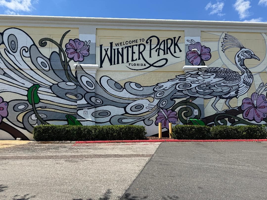 Colorful mural in Winter Park, Florida, featuring a stylized white peacock with a sweeping, detailed tail accented by abstract circular patterns and large purple hibiscus flowers. Above the bird, bold black text reads “Welcome to Winter Park, Florida,” set against a pale yellow wall under a sunny blue sky.