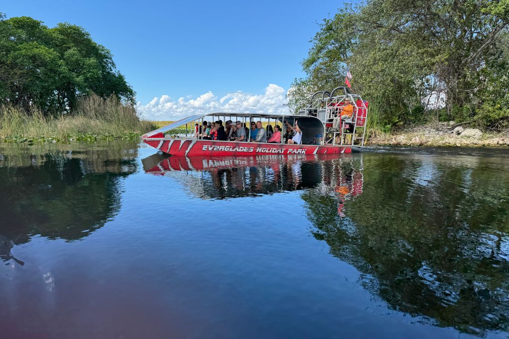 Everglades A red airboat carrying tourists glides through the calm waters of the Florida Everglades at Holiday Park, surrounded by lush greenery, tall grasses, and clear blue skies on a sunny day.