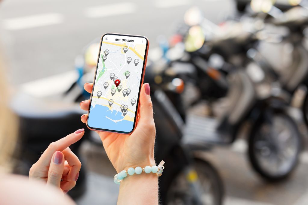 A close-up of a person using a ridesharing app on a smartphone, displaying a map with multiple vehicle location markers, while standing near a row of parked scooters or motorcycles on a city street.