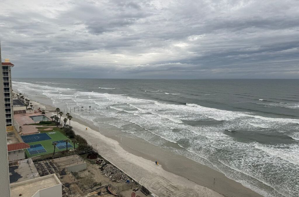 A dramatic, overcast view of Daytona Beach with gray skies and choppy waves rolling onto the wide shoreline. High-rise buildings and oceanfront resorts line the coast, while tennis and basketball courts sit near the sand. The moody atmosphere hints at a stormy day along Florida’s Atlantic Coast.