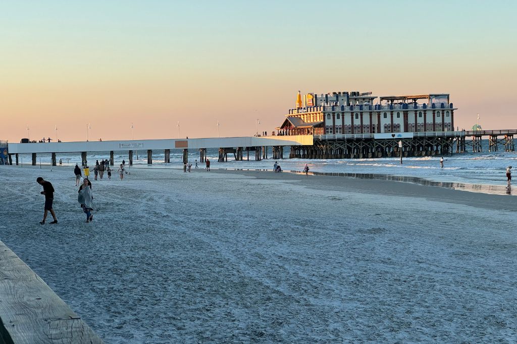 Daytona Beach People walking along the sandy shore of Daytona Beach at sunset, with the historic Daytona Beach Pier and Joe's Crab Shack extending over the ocean in the background.</p>
<p>
