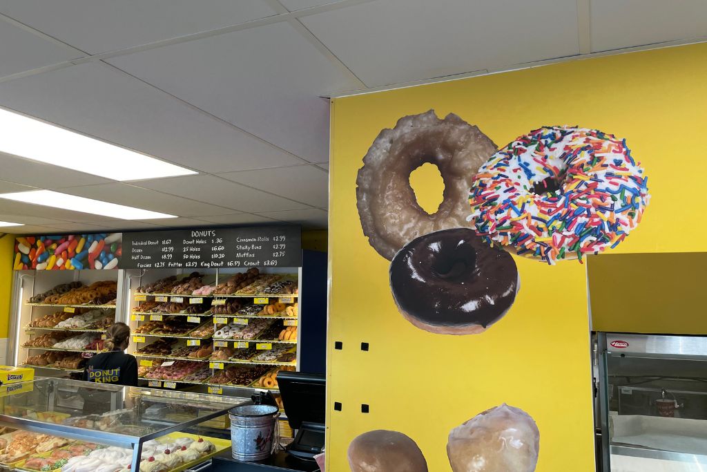 Donut King Minneola Florida Interior of a donut shop featuring colorful wall art with large images of donuts, a display case filled with pastries, and shelves stacked with a wide variety of donuts behind the counter, where a staff member is assisting customers.