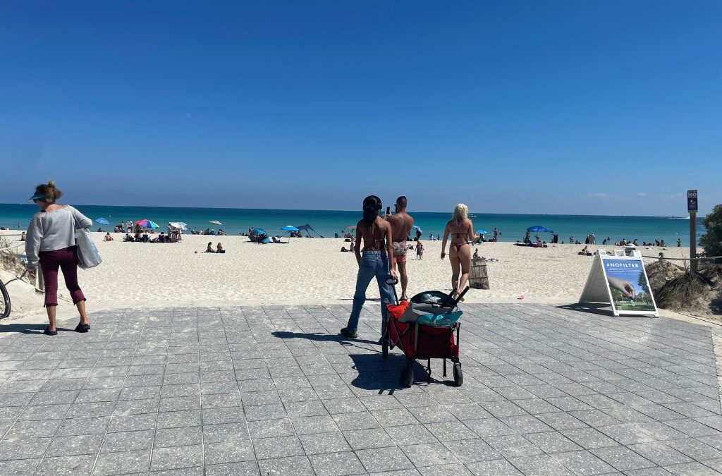Beachgoers walking toward the sandy shoreline of Miami Beach on a sunny day, with turquoise water, umbrellas, and a #NOFILTER sign displayed on the walkway.