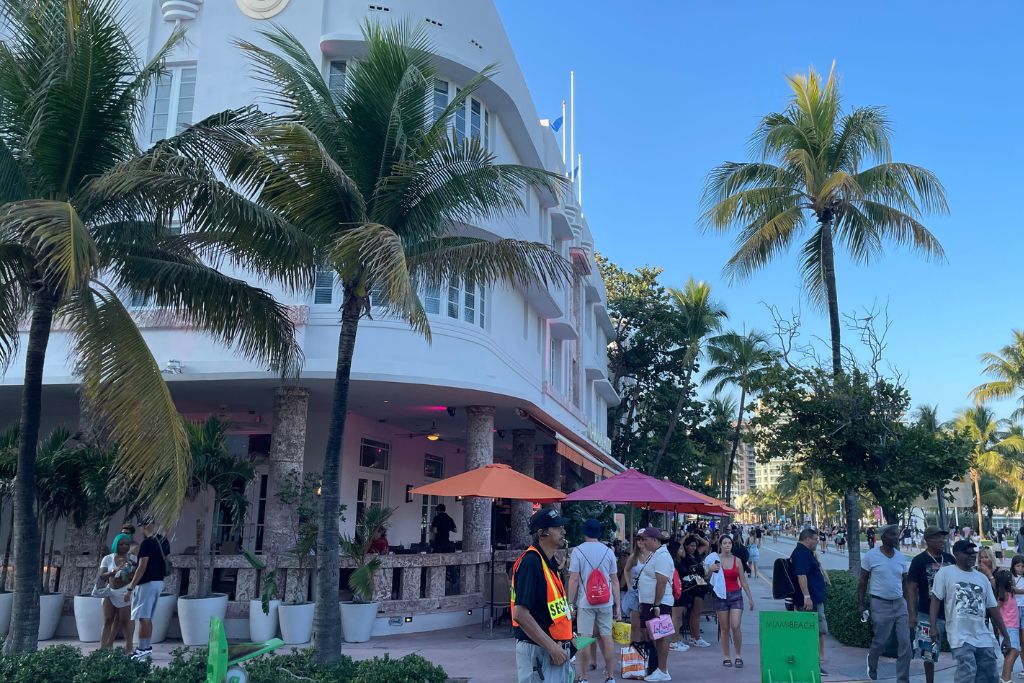 Miami Beach Crowds of people walking along Ocean Drive in Miami Beach on a sunny afternoon, with palm trees, Art Deco buildings, sidewalk cafes, and a security guard visible in the scene.