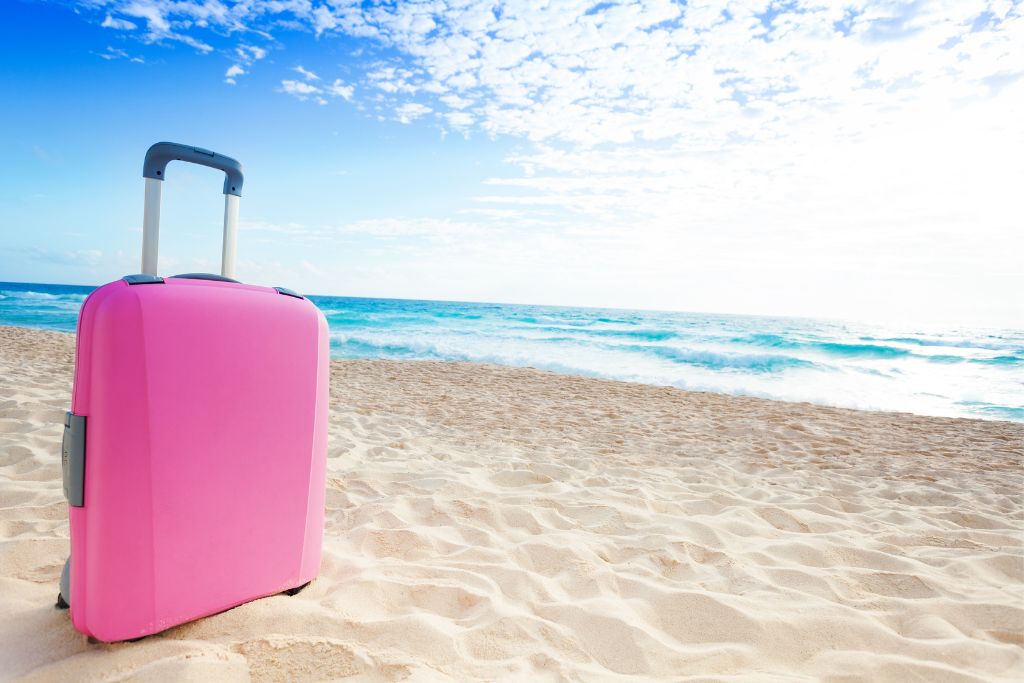 Bright pink suitcase standing on a sandy beach with turquoise waves and a blue sky in the background, symbolizing travel and vacation