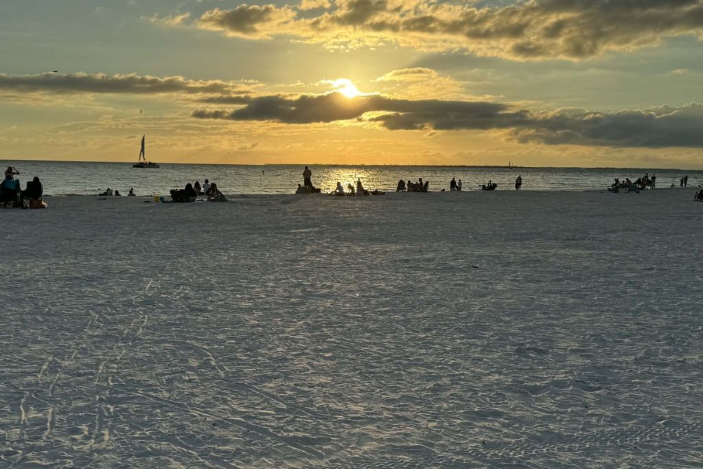 People gathered along a wide sandy beach at sunset, with the sun glowing through scattered clouds over calm ocean water and silhouettes of beachgoers watching the horizon.
