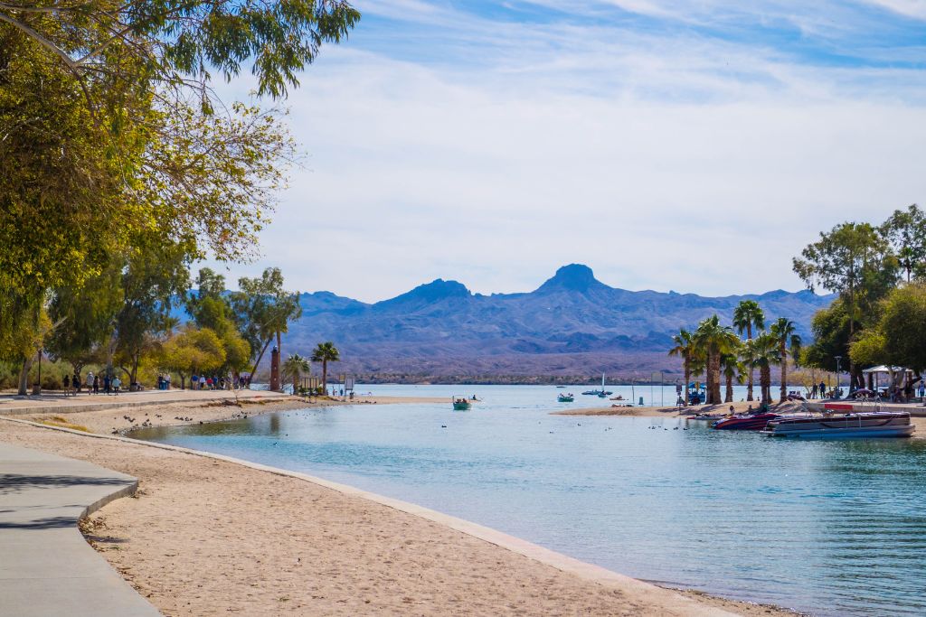 Scenic Lake Havasu City, Arizona, waterfront with sandy beaches, palm trees, boats on blue water, and rugged desert mountains in the background.