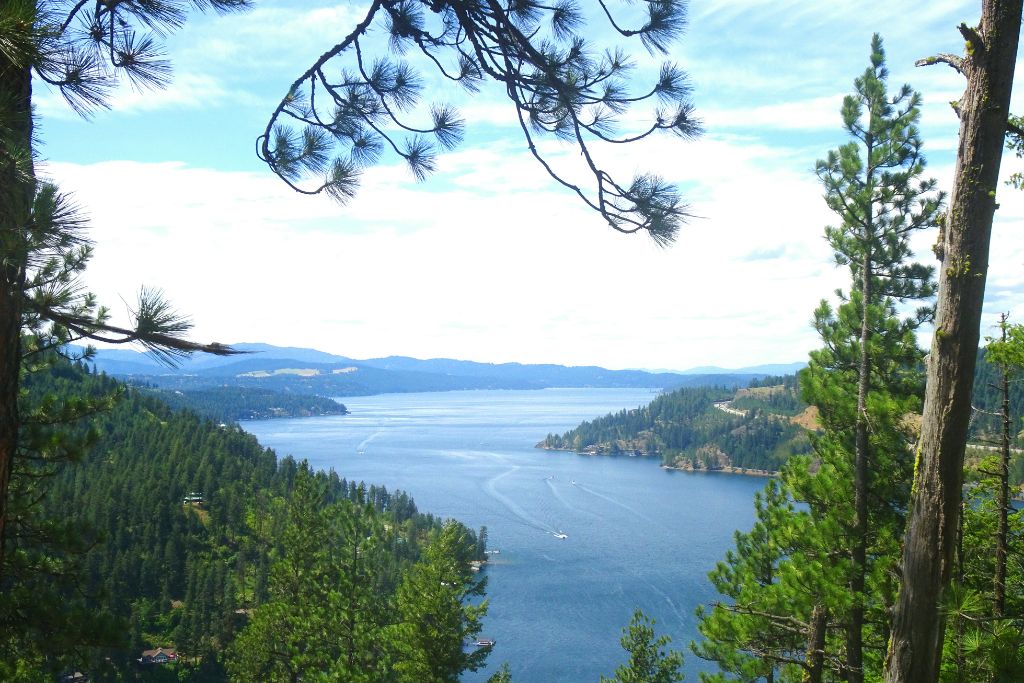 Scenic view of Lake Coeur d’Alene, Idaho, surrounded by pine-covered hills and mountains, with boats crossing the deep blue water.