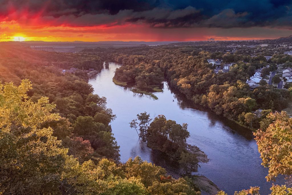 Scenic view of Branson, Missouri, with a winding river surrounded by forested hills and town neighborhoods beneath a dramatic red and orange sunset.
