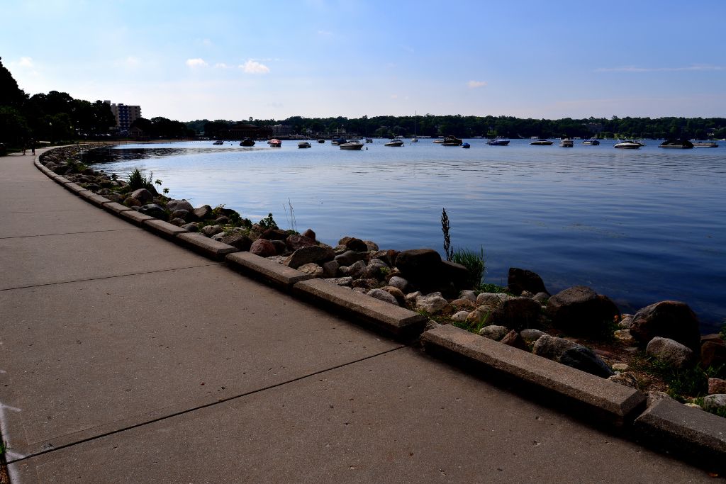 Scenic waterfront in Lake Geneva, Wisconsin, with a paved lakeside walking path, rocky shoreline, calm blue water, and boats anchored across the lake.