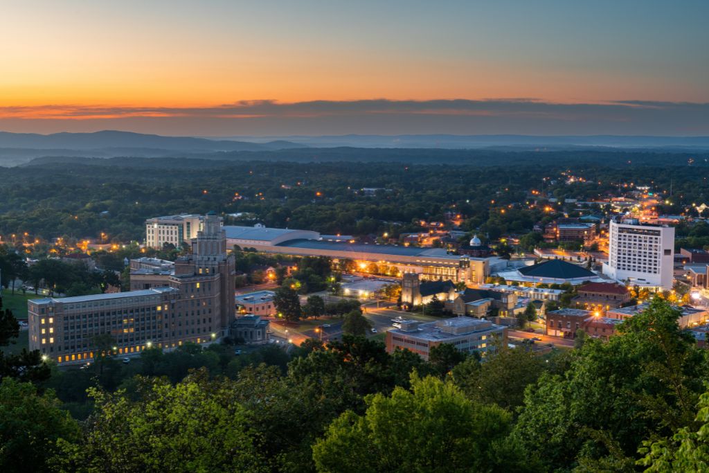 Panoramic view of downtown Hot Springs, Arkansas, illuminated at sunset, surrounded by forested hills and distant mountains.