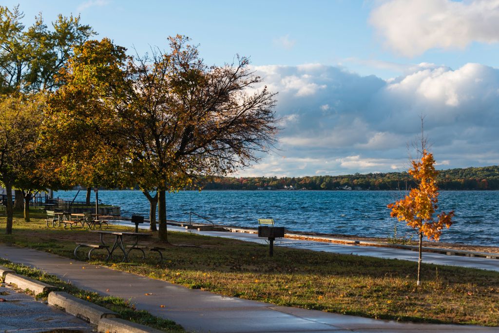 Scenic waterfront in Traverse City, Michigan, with autumn trees, picnic tables, a walking path, and blue lake water beneath a partly cloudy sky.
