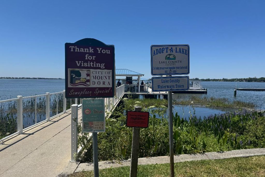 Mount Dora waterfront with Lake Dora in the background, featuring a “Thank You for Visiting City of Mount Dora” sign, Lake County Waterways Tours signage, a lakeside dock, and lush shoreline vegetation beneath a clear blue Florida sky.